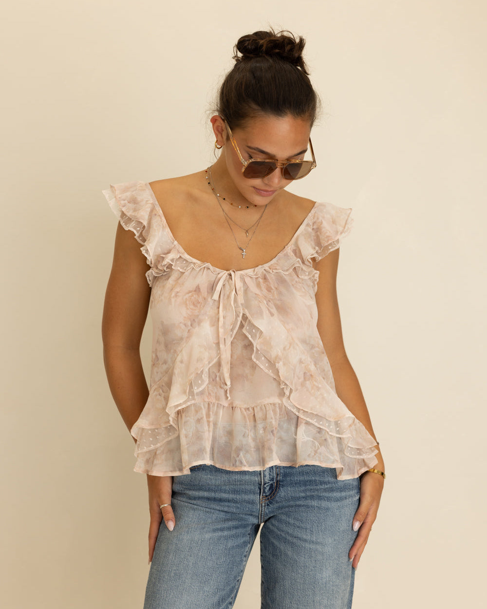 A woman wearing a light beige ruffled blouse with a subtle floral print and blue jeans, standing against a plain beige background.