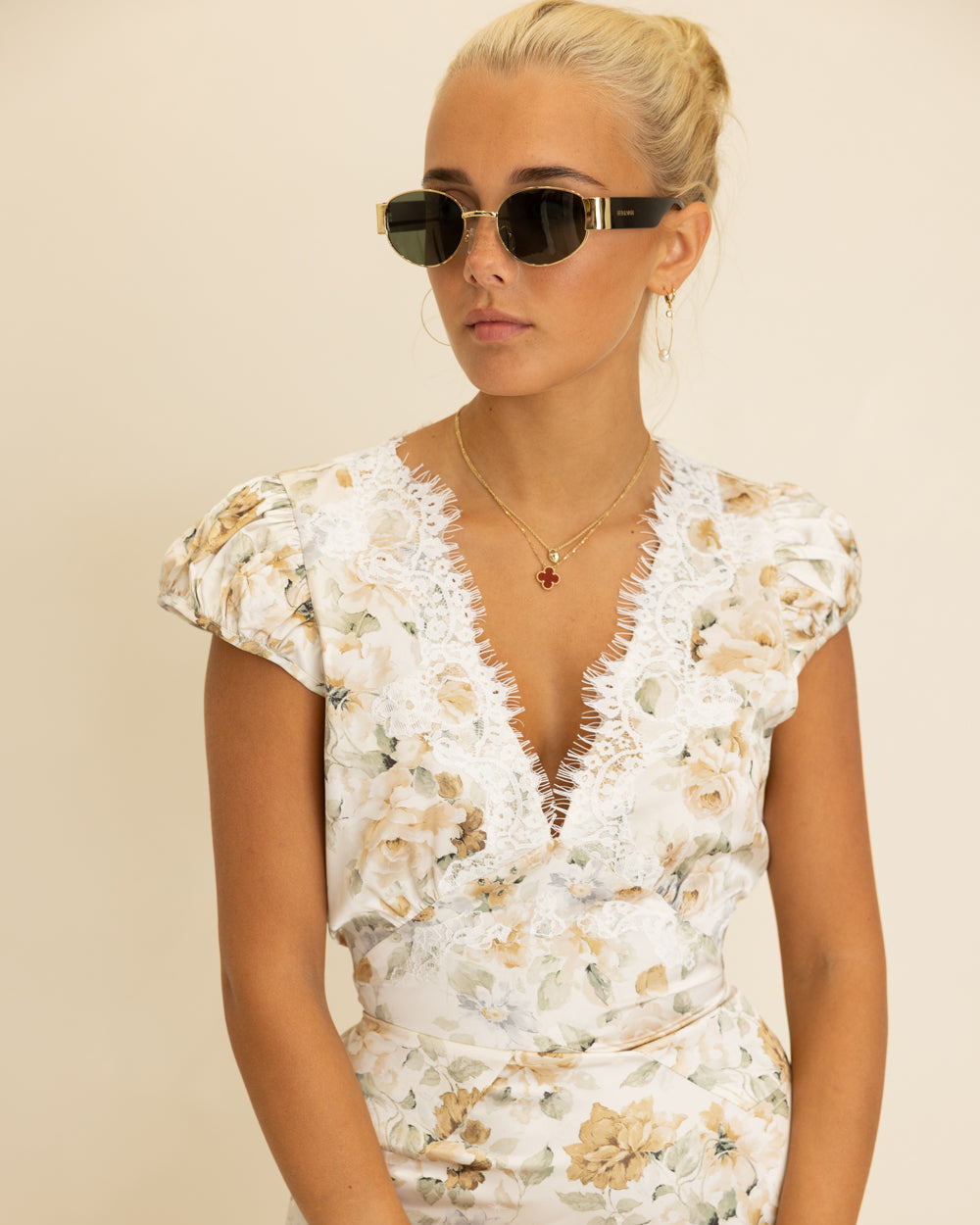 A woman wearing a white floral dress with a lace-trimmed V-neckline and cap sleeves, styled with dark sunglasses, gold necklaces, and bracelets against a plain beige background.