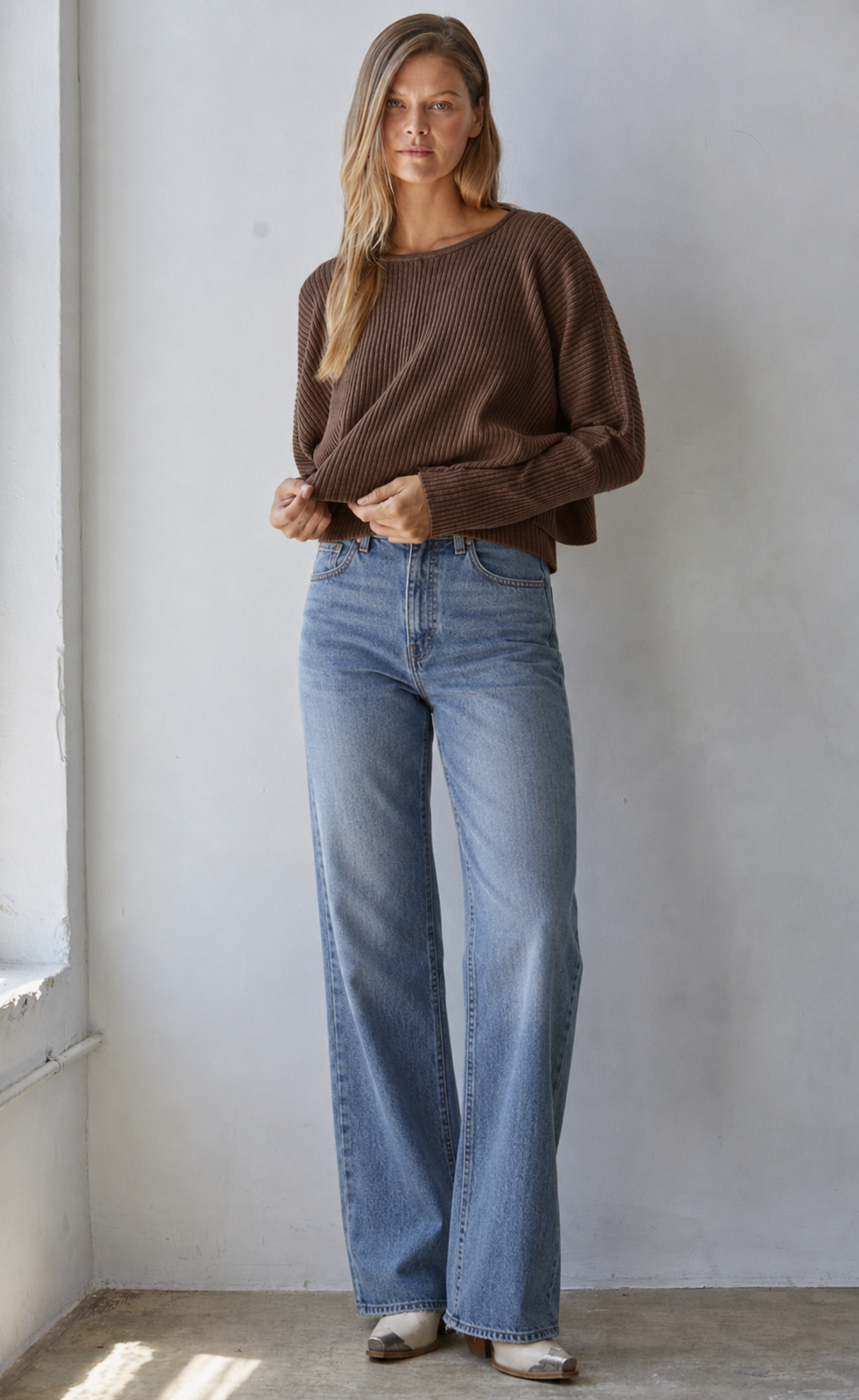 A woman stands indoors wearing a brown ribbed knit sweater and light blue wide-leg jeans, posed against a plain light gray wall.